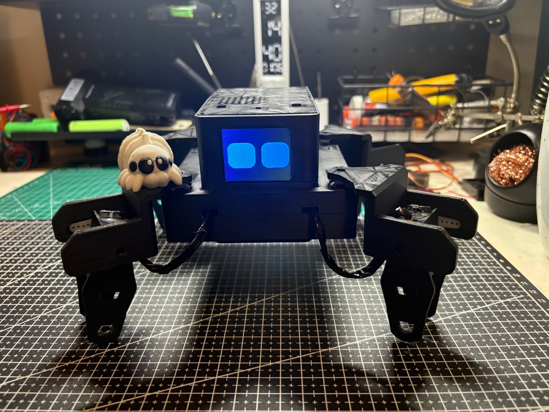 Momo, a handmade four-legged robot spider with glowing blue eyes, standing on a cutting mat at the workbench